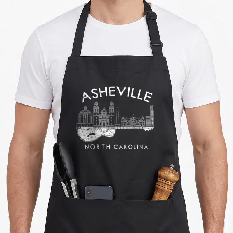 Souvenir asheville apron - White distressed Asheville skyline integrated into a guitar body shape with North Carolina — shown on a black cotton canvas apron against a white background
