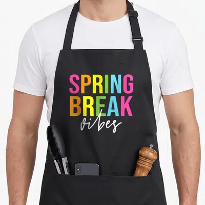 Break teacher spring aprons - Stacked SPRING BREAK vibes text in pink orange yellow green blue and white — shown on a black cotton canvas apron against a white background