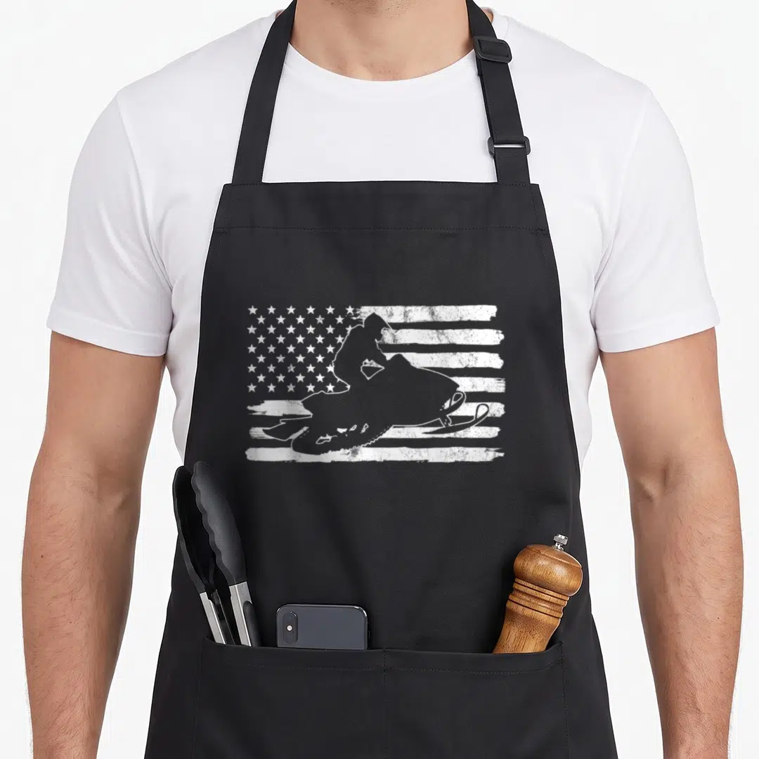 Snowmobile 4th of july apron - White distressed US flag with superimposed snowmobile rider silhouette — shown on a black cotton canvas apron against a white background