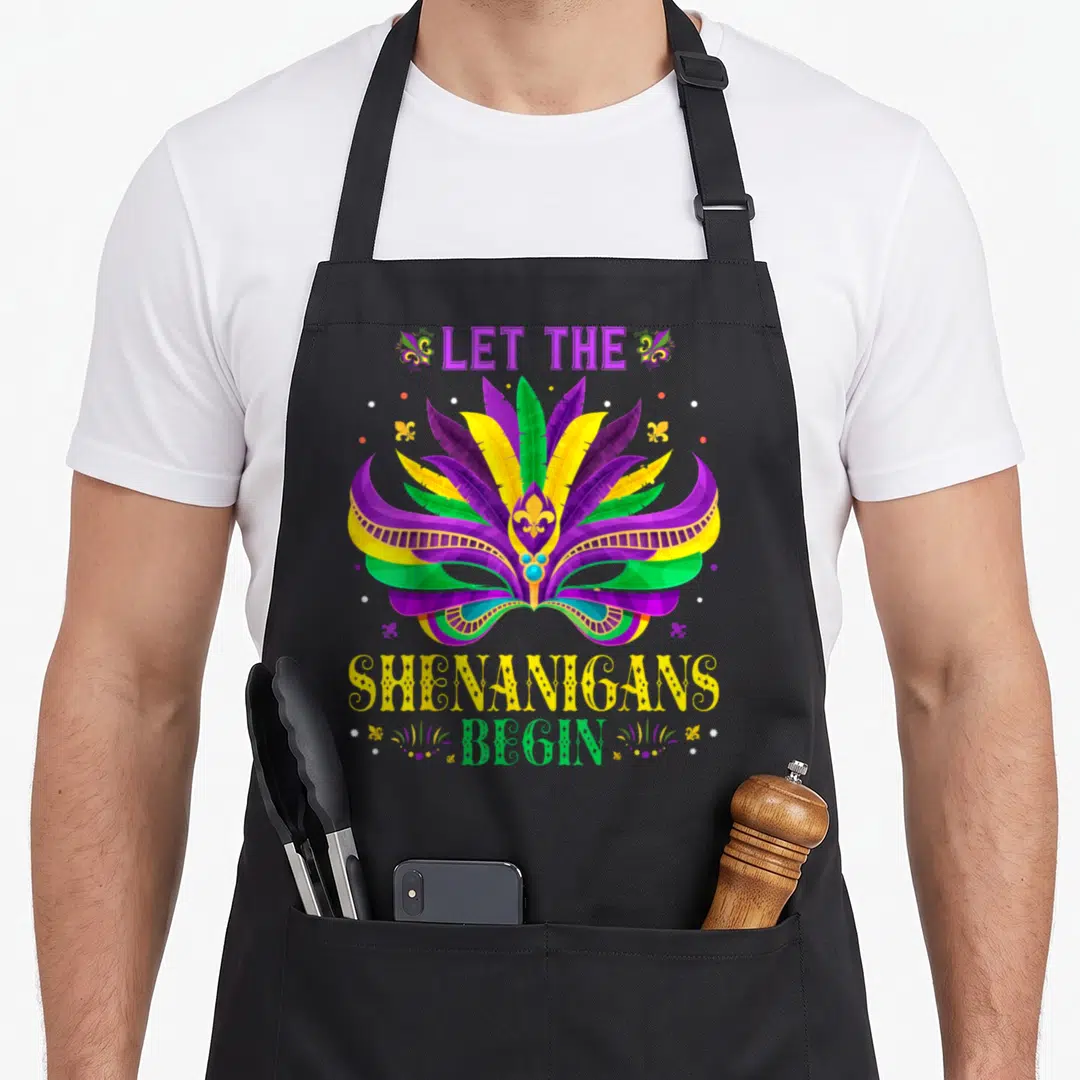 Tuesday fat chef apron - Masquerade mask in purple green yellow with LET THE SHENANIGANS BEGIN text and fleur de lis — shown on a black cotton canvas apron against a white background