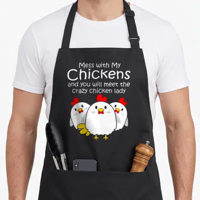 Backyard chicken with apron - Four lines of text above three cartoon white chickens with red combs and yellow beaks — shown on a black cotton canvas apron against a white background