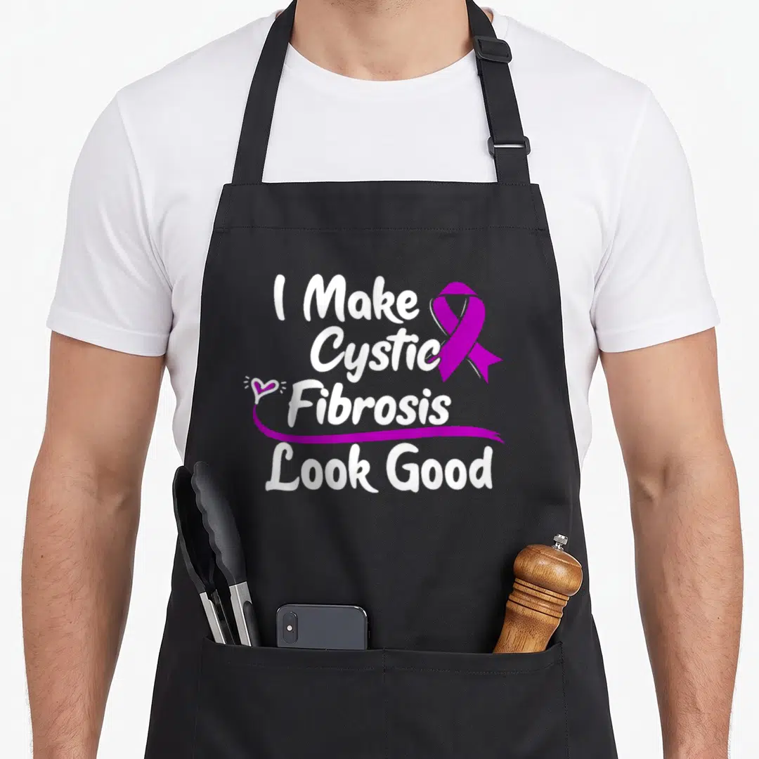 Survivor cystic fibrosis awareness apron - White stacked text I Make Cystic Fibrosis Look Good with purple ribbon — shown on a black cotton canvas apron against a white background