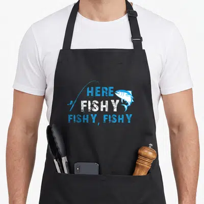 Fisherman fishing apron - Here Fishy Fishy Fishy text with blue fish and rod illustration — shown on a black cotton canvas apron against a white background