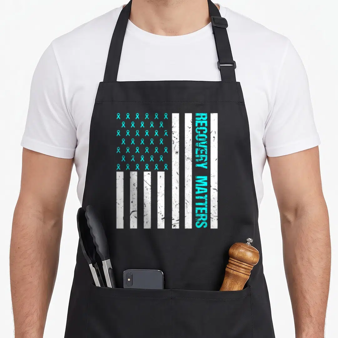 Matter addiction recovery apron - Turquoise ribbons and vertical RECOVERY MATTERS text on distressed white flag stripes — shown on a black cotton canvas apron against a white background