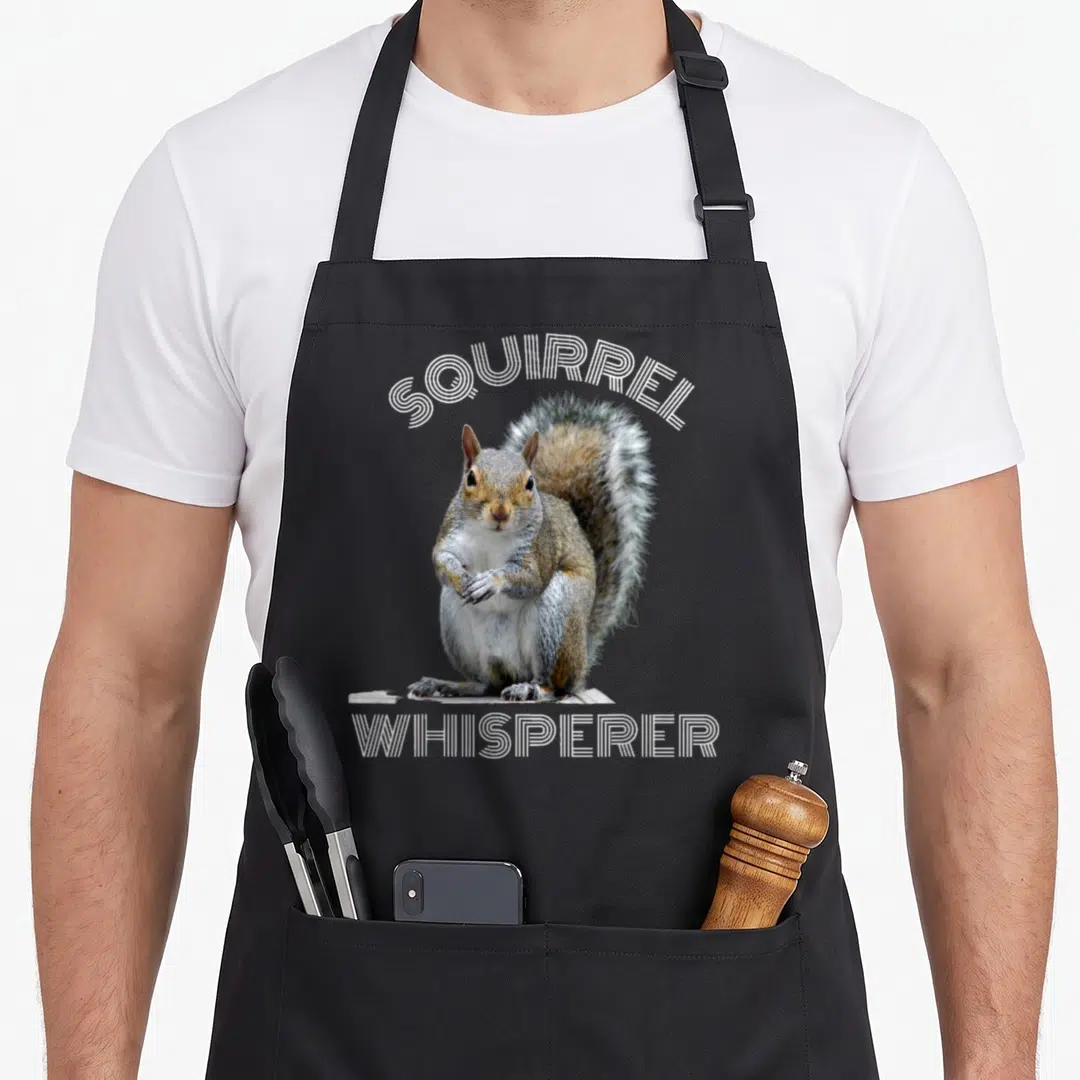 Sarcastic squirrel aprons - Photo of a grey and brown squirrel sitting on wood with arched SQUIRREL WHISPERER text — shown on a black cotton canvas apron against a white background