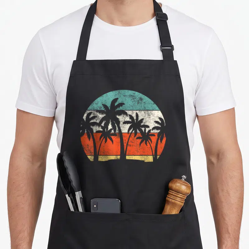Coconut holiday apron - Six grey palm tree silhouettes over circular sun with teal cream orange yellow horizontal — shown on a black cotton canvas apron against a white background