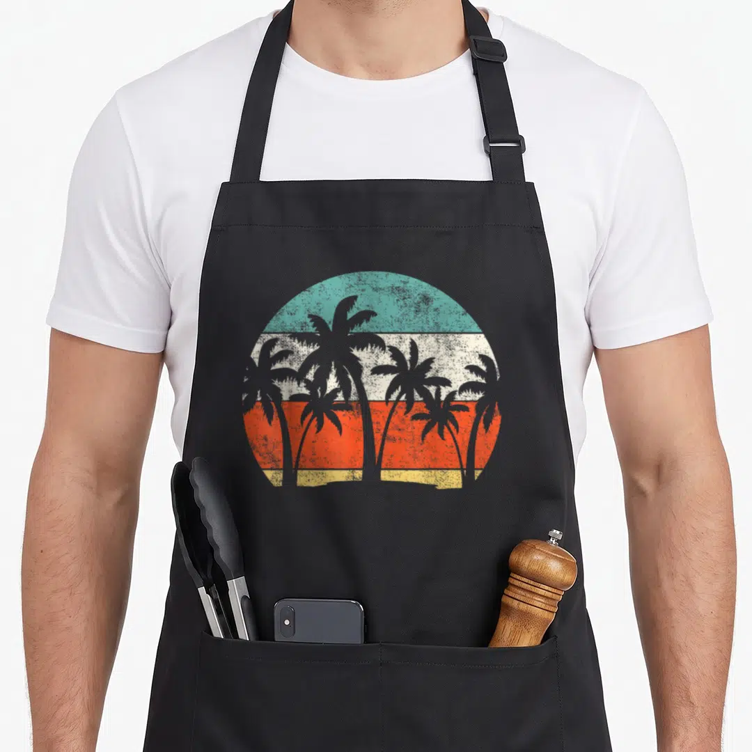 Coconut holiday apron - Six grey palm tree silhouettes over circular sun with teal cream orange yellow horizontal — shown on a black cotton canvas apron against a white background