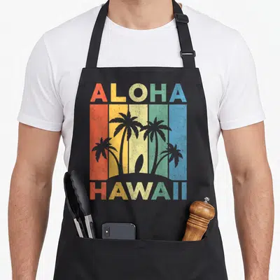 Hawaii kitchen island apron - Retro ALOHA HAWAII text with palm tree silhouettes over five colored vertical stripes — shown on a black cotton canvas apron against a white background
