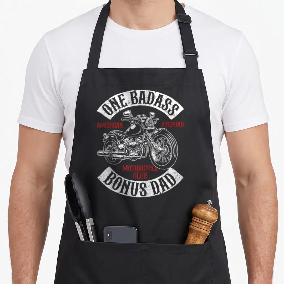Biker badass apron - White distressed motorcycle with One Badass Bonus Dad arched banners and red American Stepdad text — shown on a black cotton canvas apron against a white background