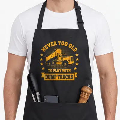 Play dump truck apron - Orange dump truck with text NEVER TOO OLD TO PLAY WITH DUMP TRUCKS and two columns of five stars — shown on a black cotton canvas apron against a white background