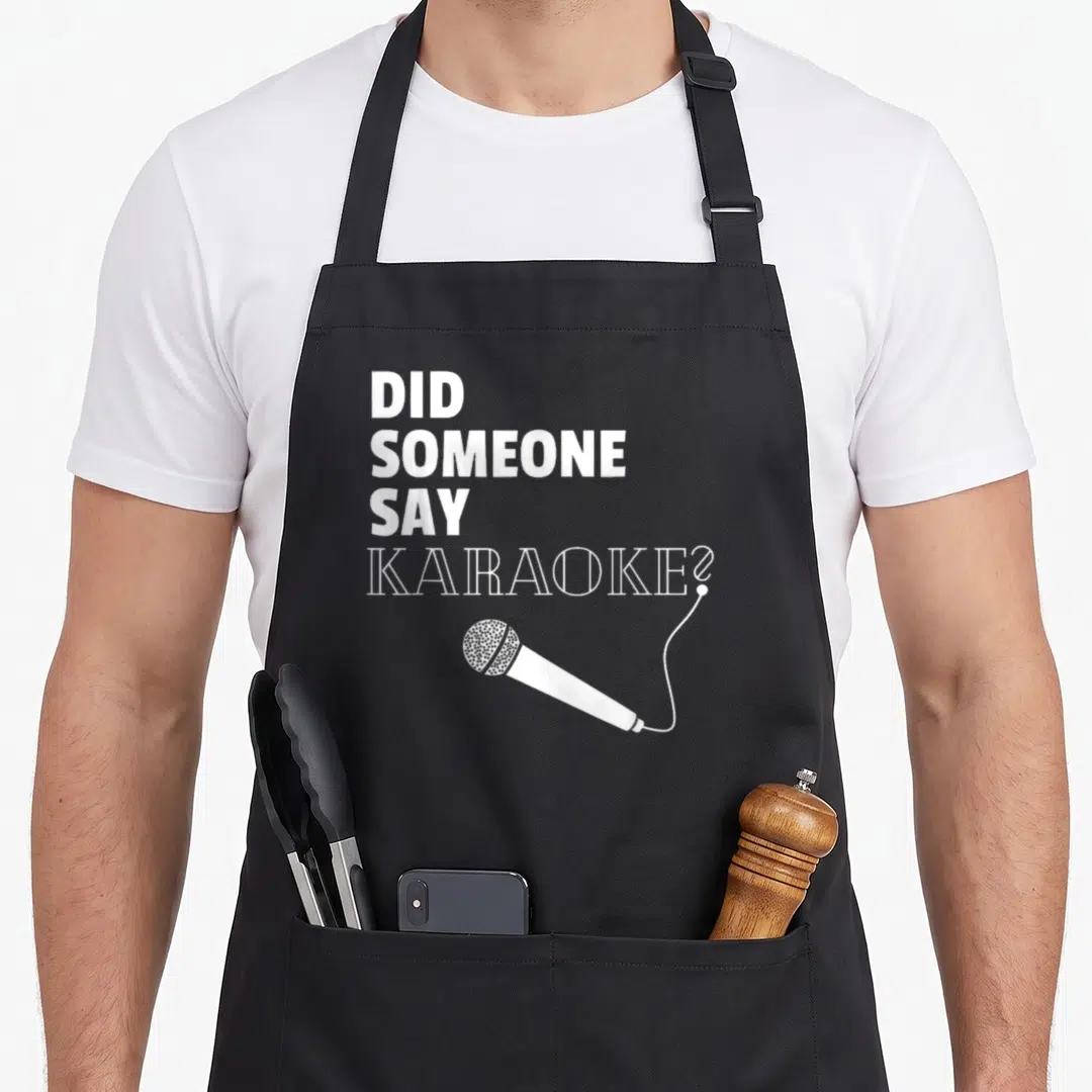 Someone kare apron - White stacked text Did Someone Say Karaoke above a diagonal microphone with a dotted mesh head — shown on a black cotton canvas apron against a white background