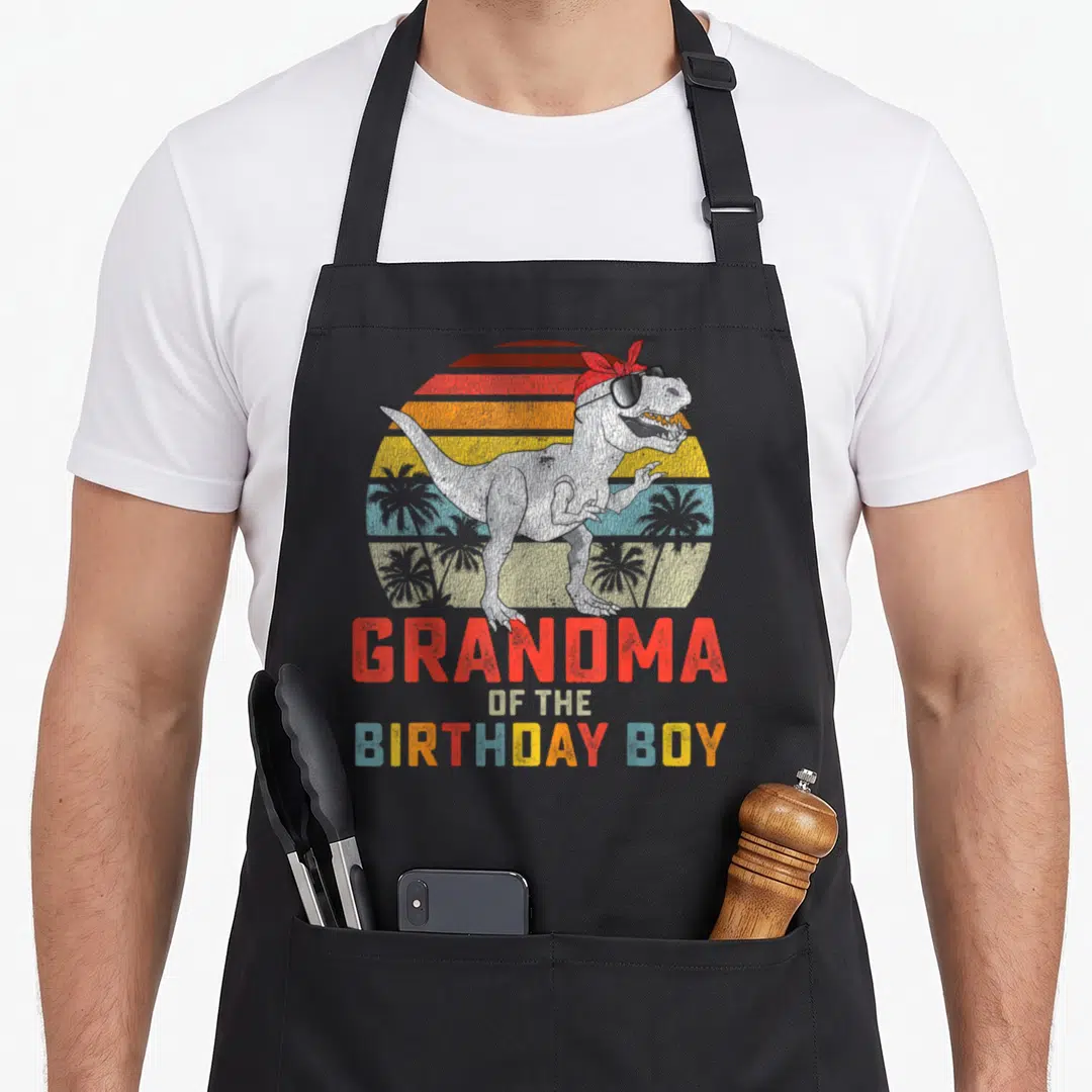 Dinosaur grandma in apron - Retro T-Rex in sunglasses and bandana with stacked GRANDMA OF THE BIRTHDAY BOY text — shown on a black cotton canvas apron against a white background