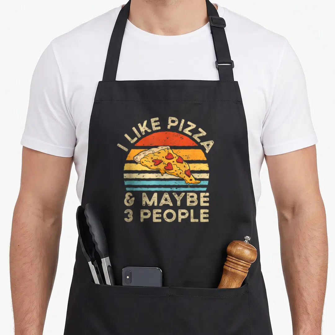 Maybe pizza apron - Retro sun with striped bands, pizza slice with heart toppings, and beige arched text reading I LIKE — shown on a black cotton canvas apron against a white background