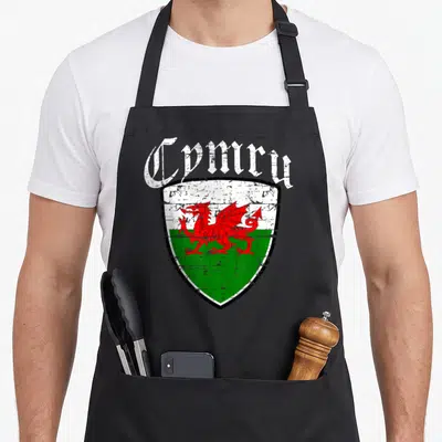 Cymru welsh apron - Distressed red dragon on white and green shield with arched Cymru text — shown on a black cotton canvas apron against a white background
