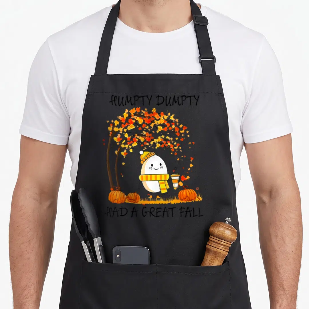 Egg cute fall aprons - Humpty Dumpty text above egg character with scarf and coffee under orange tree leaves — shown on a black cotton canvas apron against a white background