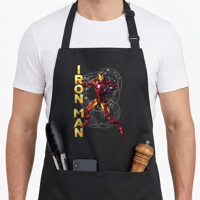 Assemble ironman apron - Vertical gold IRON MAN text beside red and gold armored figure with white circuitry background — shown on a black cotton canvas apron against a white background