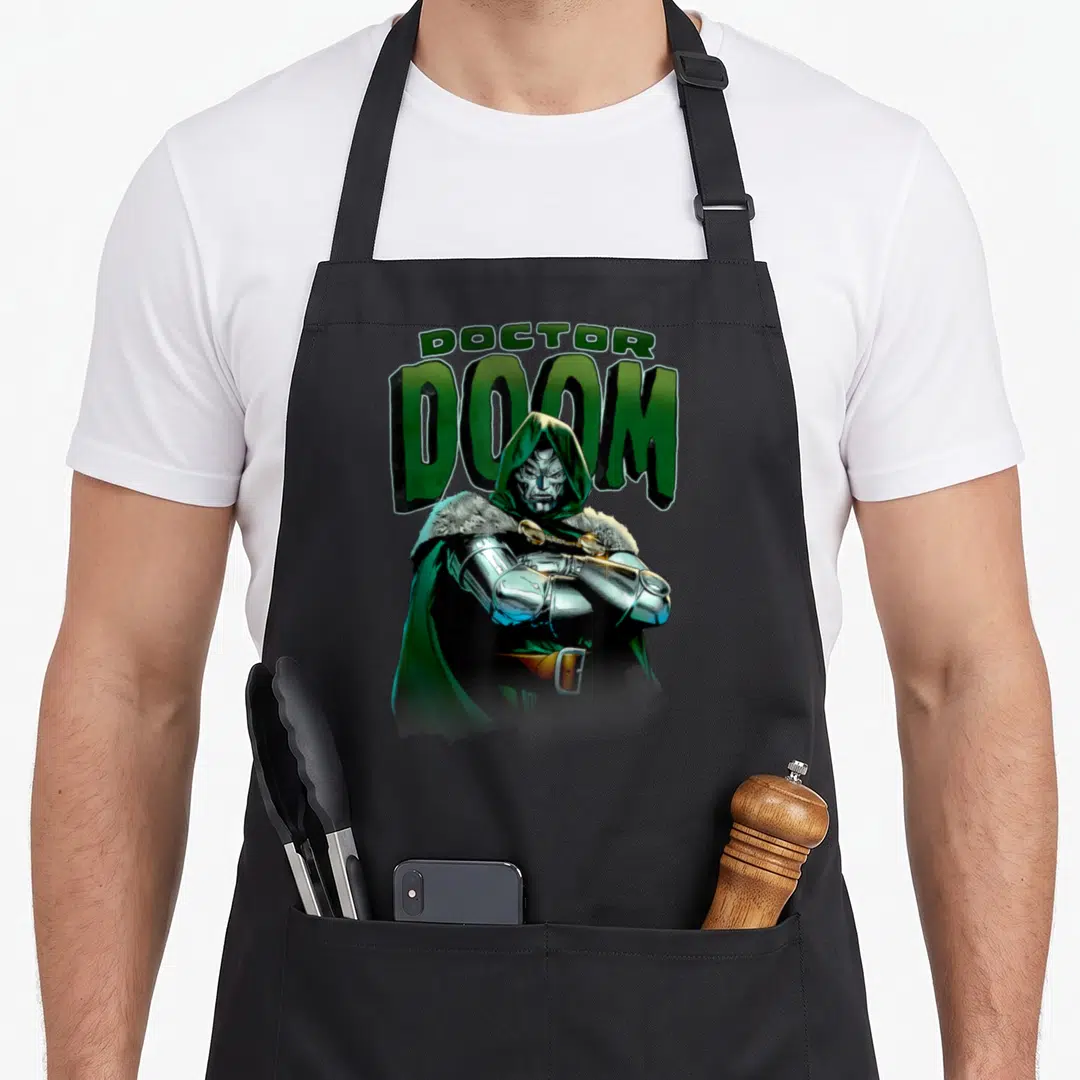 Crossed doctor apron - Stacked DOCTOR DOOM text above hooded figure with crossed metallic arms in green and silver — shown on a black cotton canvas apron against a white background