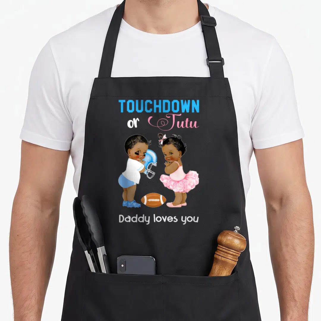 Ethnic daddy apron - Two cartoon babies with football helmet and pink tutu flanking a brown football under blue — shown on a black cotton canvas apron against a white background