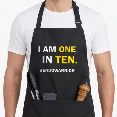 Funny endometriosis awareness apron - White and yellow text reading I AM ONE IN TEN and #ENDOWARRIOR stacked vertically — shown on a black cotton canvas apron against a white background