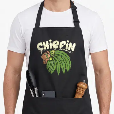 Chiefin smoking apron - Arched CHIEFIN text above Native American chief profile with green headdress in distressed — shown on a black cotton canvas apron against a white background