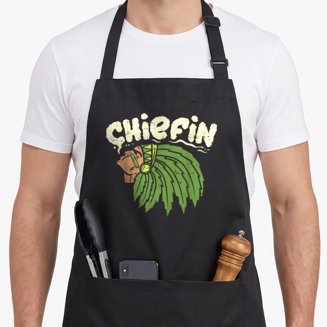 Chiefin smoking apron - Arched CHIEFIN text above Native American chief profile with green headdress in distressed — shown on a black cotton canvas apron against a white background