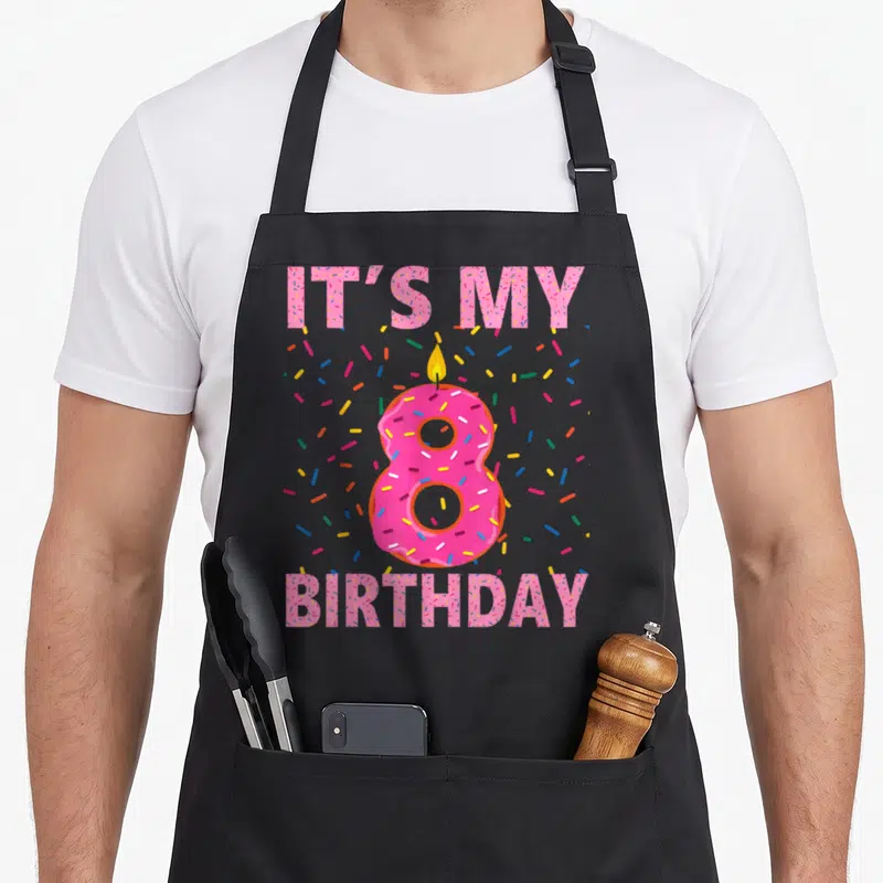 Sweet donut apron - Pink number 8 donut with candle and IT'S MY BIRTHDAY text surrounded by sprinkles — shown on a black cotton canvas apron against a white background