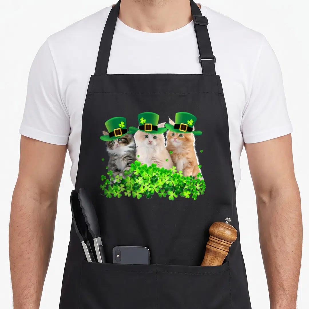 Three st patrick day aprons - Three cats in green hats behind a cluster of green shamrocks — shown on a black cotton canvas apron against a white background