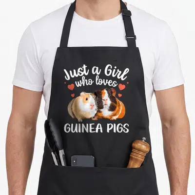 Clothe guinea lover apron - Two cartoon guinea pigs in white brown orange and red with scattered red hearts and printed — shown on a black cotton canvas apron against a white background