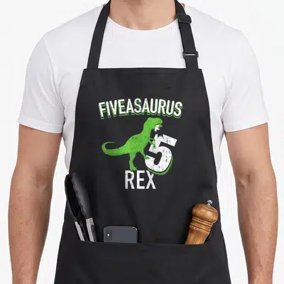 Dino t-rex apron - Green cartoon dinosaur leaning on white number 5 under arched FIVEASAURUS text — shown on a black cotton canvas apron against a white background