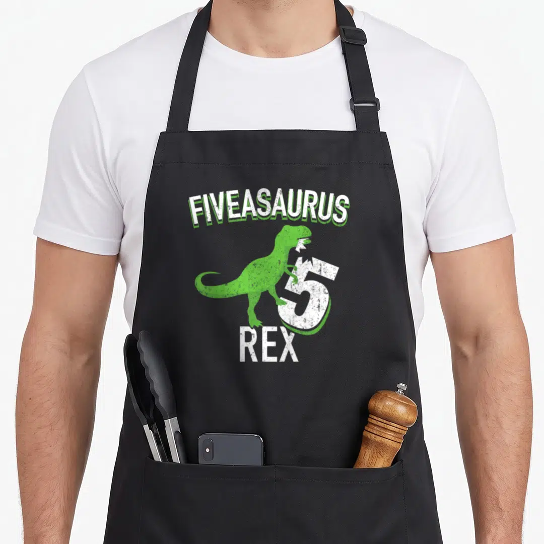 Dino t-rex apron - Green cartoon dinosaur leaning on white number 5 under arched FIVEASAURUS text — shown on a black cotton canvas apron against a white background