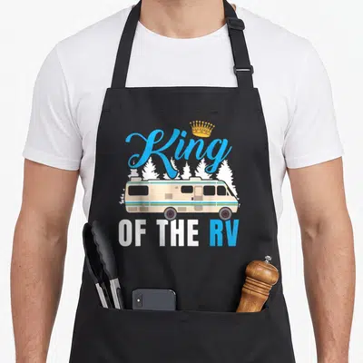 Owner king apron - Beige RV illustration with blue King OF THE RV text and golden crown graphic — shown on a black cotton canvas apron against a white background