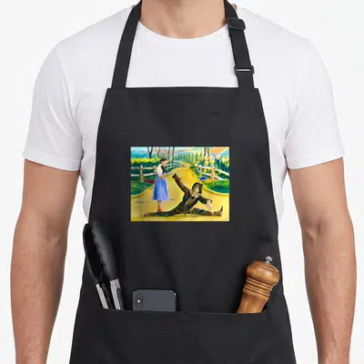 Land dorothy apron - Photo of woman in blue dress helping reclining scarecrow on yellow road with green hills — shown on a black cotton canvas apron against a white background