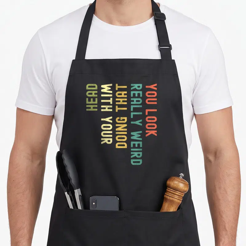 Funny doing weird aprons - Vertical stacked text reading You Look Really Weird Doing That With Your Head in orange teal — shown on a black cotton canvas apron against a white background