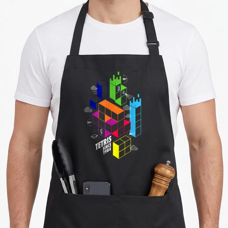 Play gamer apron - Isometric tetris blocks in blue green orange red yellow and white with TETRIS SINCE 1984 text — shown on a black cotton canvas apron against a white background