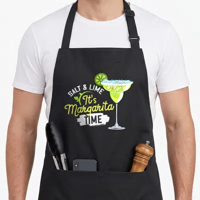 Margarita limes apron - Cartoon margarita glass with ice and lime slice next to stacked text reading SALT & LIME It's — shown on a black cotton canvas apron against a white background