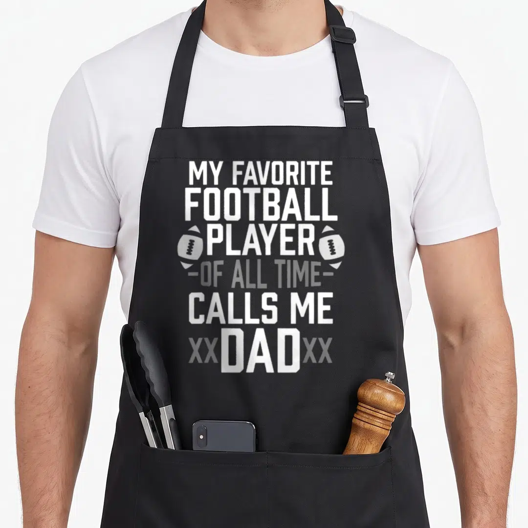 Call football apron - White stacked text My Favorite Football Player Calls Me Dad with two football icons on black — shown on a black cotton canvas apron against a white background