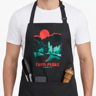 Tonal twin peaks apron - Retro landscape with red sun, grey mountains, teal trees, and text TWIN PEAKS population 51,201 — shown on a black cotton canvas apron against a white background