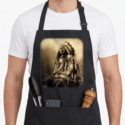 Funny chiefs apron - Sepia photo of seated Native American man in traditional dress holding a spear inside rounded frame — shown on a black cotton canvas apron against a white background