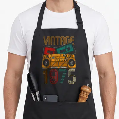 Imported birthday apron - Orange boombox with red and teal cassettes and VINTAGE 1975 text — shown on a black cotton canvas apron against a white background