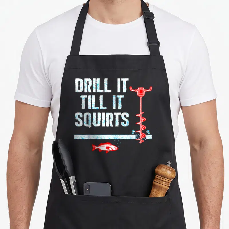 Drill fishing aprons - Distressed white text DRILL IT TILL IT SQUIRTS beside red ice auger and red fish under white line — shown on a black cotton canvas apron against a white background