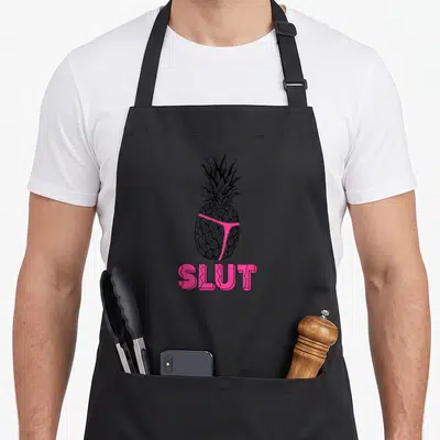 Caption brooklyn apron - Black and white pineapple illustration wearing a pink thong above the word SLUT in pink block — shown on a black cotton canvas apron against a white background