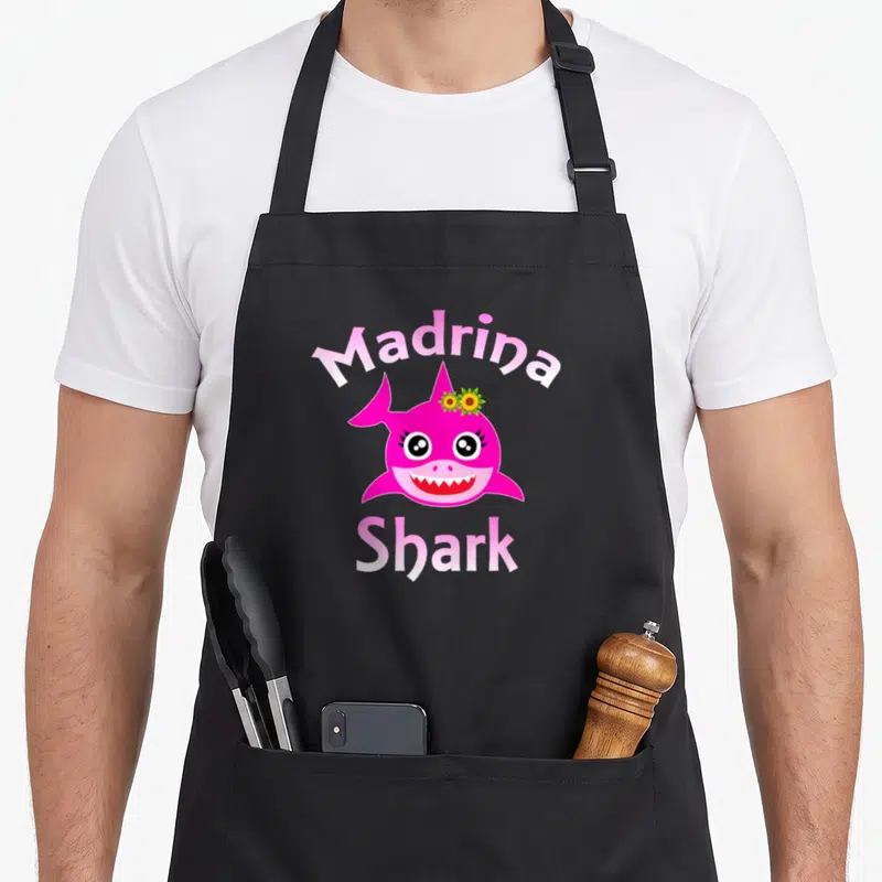 Madrina spanish aprons - Pink cartoon shark head with two yellow sunflowers and Madrina Shark text — shown on a black cotton canvas apron against a white background