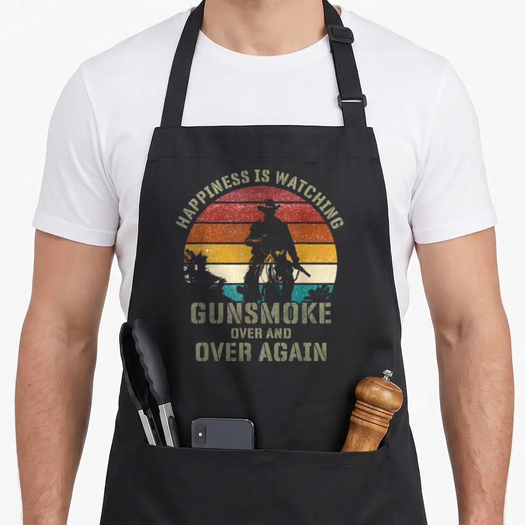 Happiness cowboy apron - Distressed retro sun with cowboy silhouette and Gunsmoke quote text — shown on a black cotton canvas apron against a white background