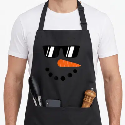 Toddler snowman apron - Minimalist snowman face with black sunglasses, orange carrot nose, and curved row of black — shown on a black cotton canvas apron against a white background