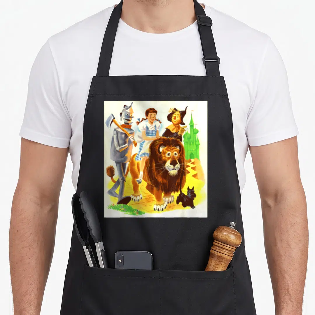 Cowardly dorothy apron - Dorothy riding lion on yellow brick road with Tin Man Scarecrow Toto green castle — shown on a black cotton canvas apron against a white background