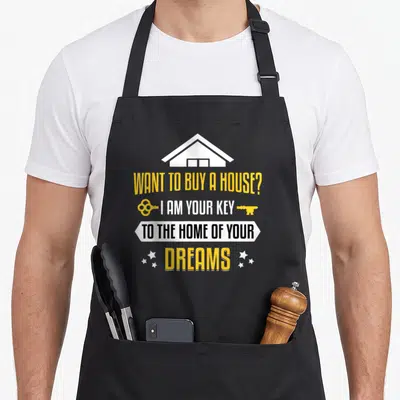 Key real estate agent apron - House icon and WANT TO BUY A HOUSE text with two keys and stars in yellow white black — shown on a black cotton canvas apron against a white background
