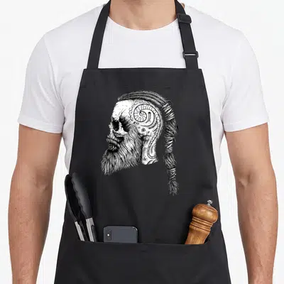 Nordic country apron - Black and white line art skull with beard tattoos and braided ponytail — shown on a black cotton canvas apron against a white background