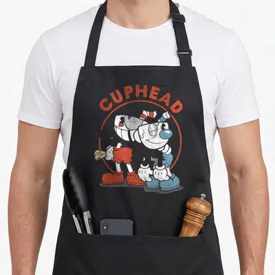 Cuphead beat apron - Cuphead and Mugman cartoon characters under arched CUPHEAD text with Also Starring MUGMAN caption — shown on a black cotton canvas apron against a white background