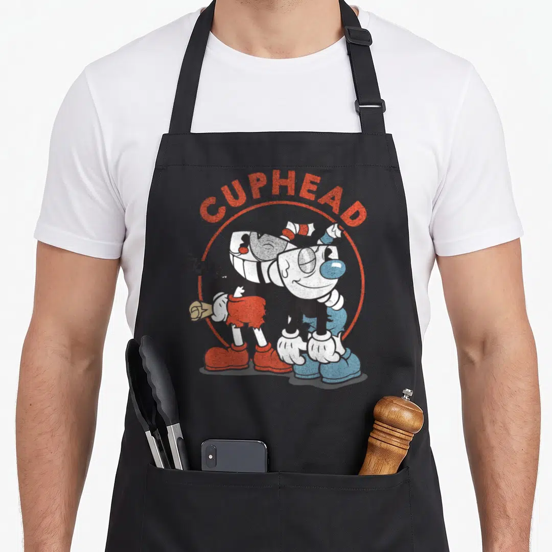 Cuphead beat apron - Cuphead and Mugman cartoon characters under arched CUPHEAD text with Also Starring MUGMAN caption — shown on a black cotton canvas apron against a white background