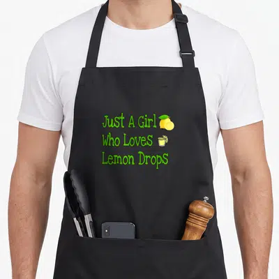 Cute drop for love and lemons apron - Green text Just A Girl Who Loves Lemon Drops with yellow lemon and cocktail glass — shown on a black cotton canvas apron against a white background